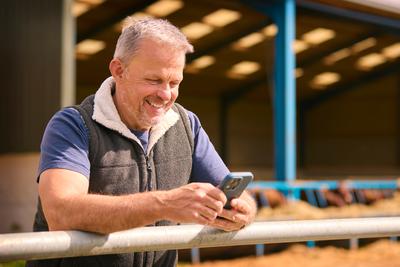 A farmer smiling while checking his phone, leaning against a gate outside a farm building